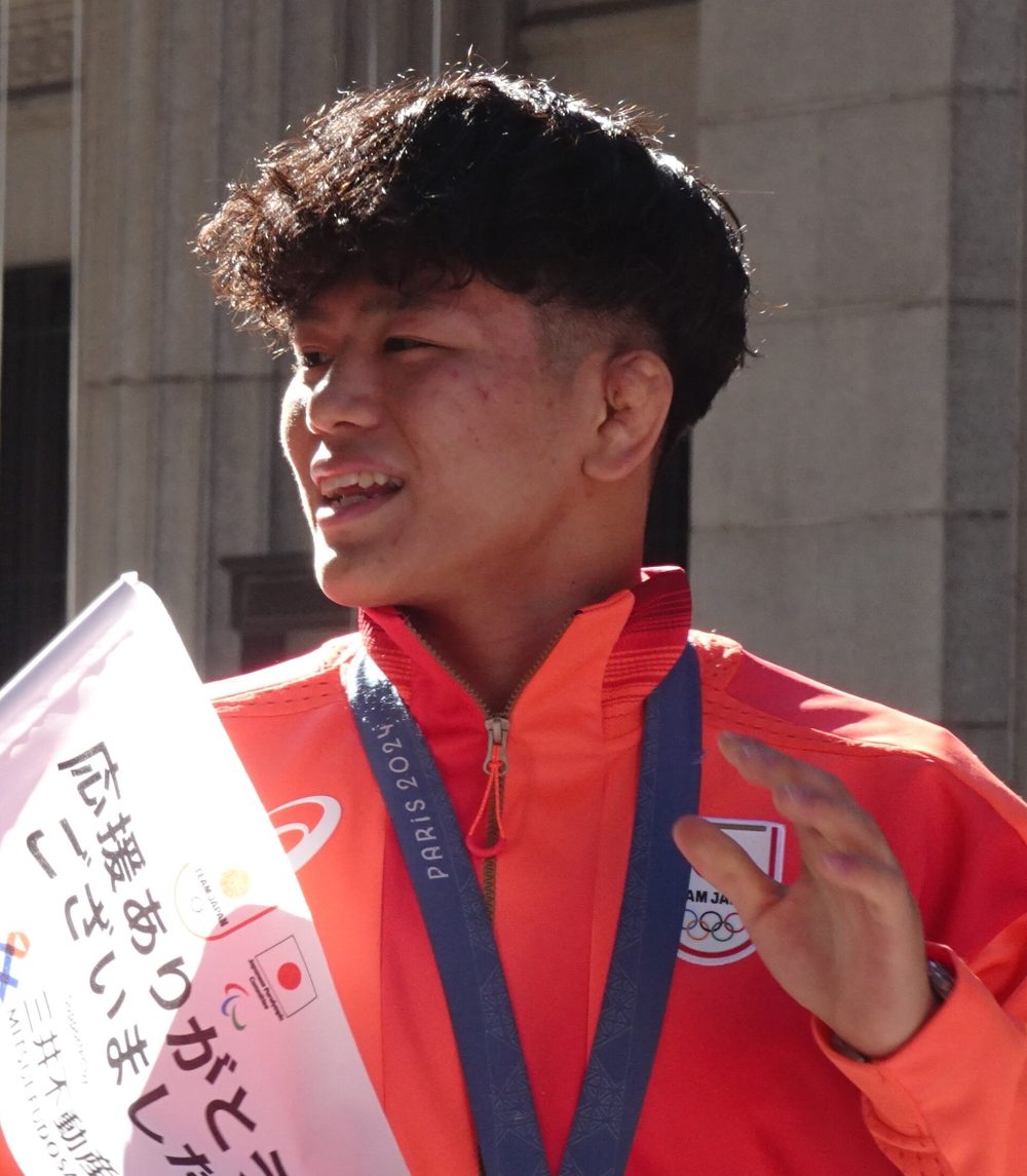 Close portrait of Kotaro Kiyooka in a Team Japan jacket with his gold medal ribbon, holding a thank-you flag